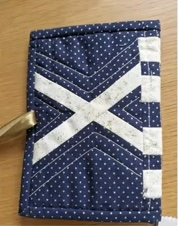 A blue quilted needle case with a white "X" shape in the center. The background fabric has a small polka dot pattern.
