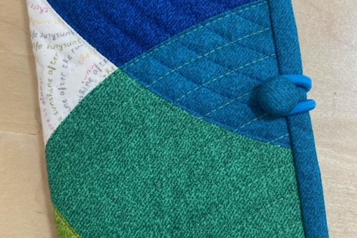 A blue quilted needle case with a patchwork design. The patchwork features shades of blue, green, and white fabric, arranged in a curved pattern. The case has a blue button closure.