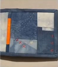 A blue quilted needle case with a patchwork design. The patchwork features various shades of blue, white, and a bright orange stripe. There are also small embroidered crosses and snowflakes on the case.
