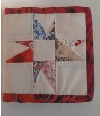 A red quilted needle container with a patchwork design. The patchwork features a star shape made up of red, white, and blue fabric pieces. The container has a red border.