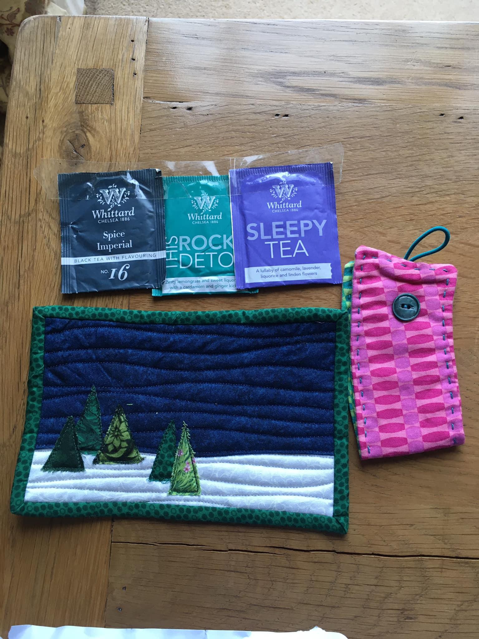 A mug rug with Christmas trees in the snow and a dark blue sky. The mugrug is bound with green spotty fabric. To the right of the mugrug is a mug cozy made of a pink patterned fabric with green big hand-stitching and a green button with elastic closure. Above the mugrug are some sachets of tea.