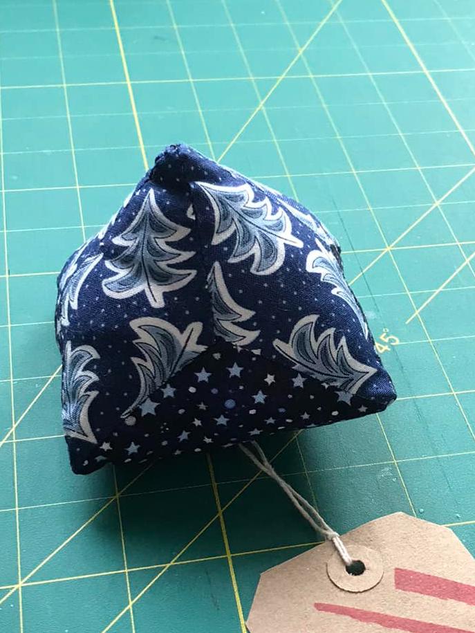 An triangular shaped pincushion with sides made of a blue fabric with Christmas trees. The front edge has stars and spots. A tag is attached to the pincushion.