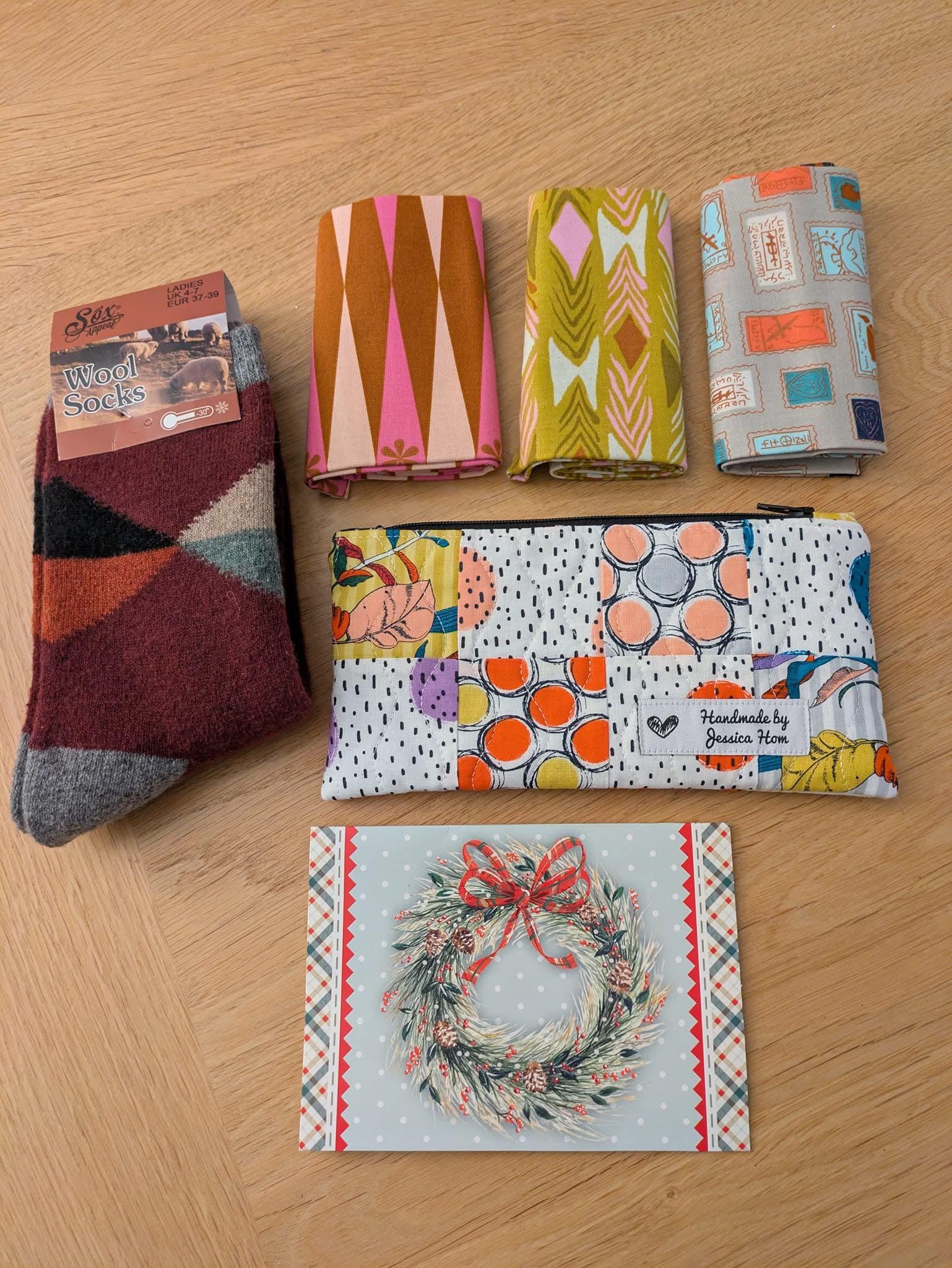 A patchwork zippered pouch made of white and coloured patterned squares and a black zip. There are 3 lots of folded fabric above the pouch, a pair of patchwork inspired socks to the left and a Christmas card below.