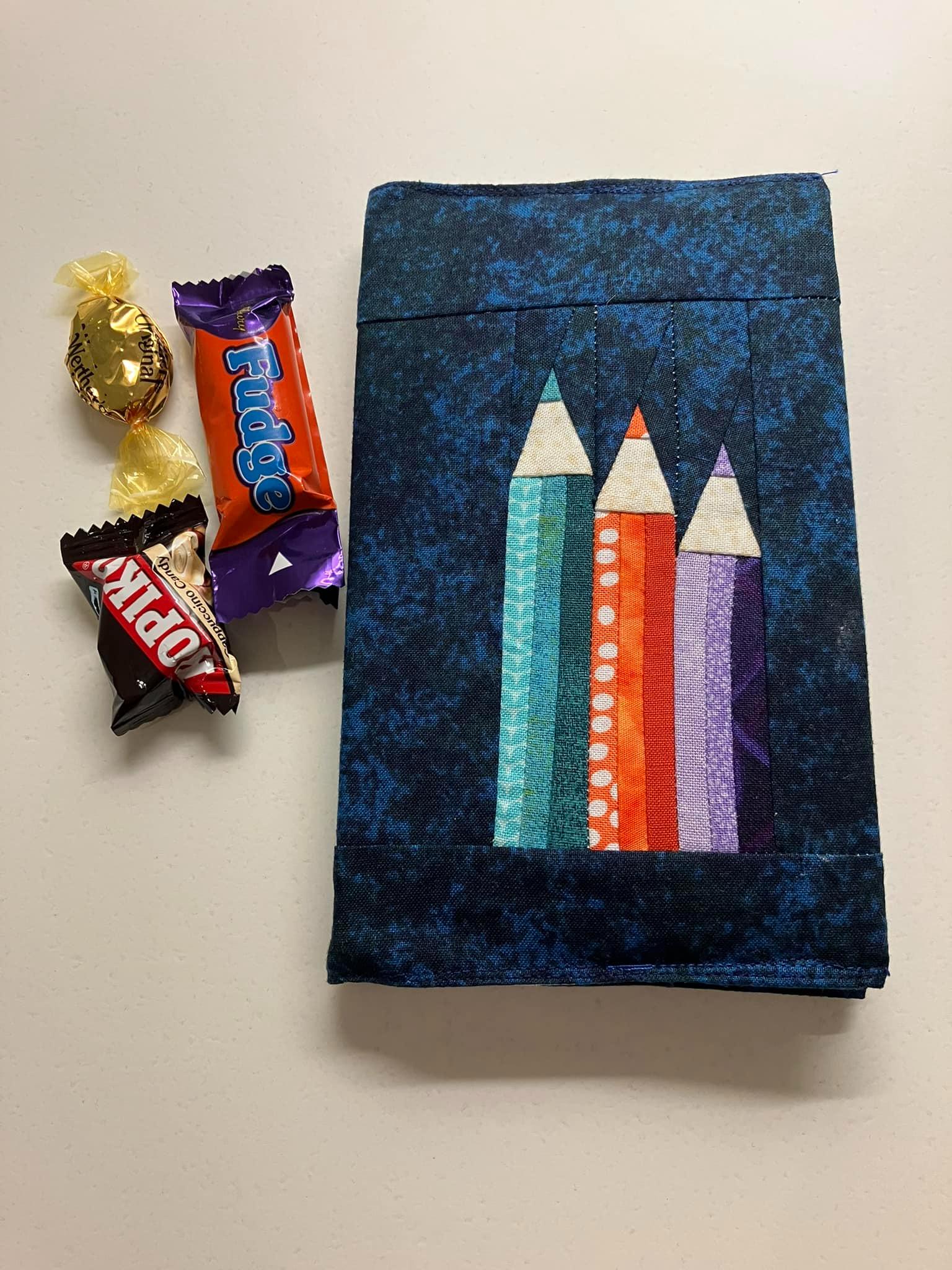 A notebook cover with a navy background and a pieced trio of pencils in descending height as the focal point. Some selection box sized chocolates lie to the left of the cover.