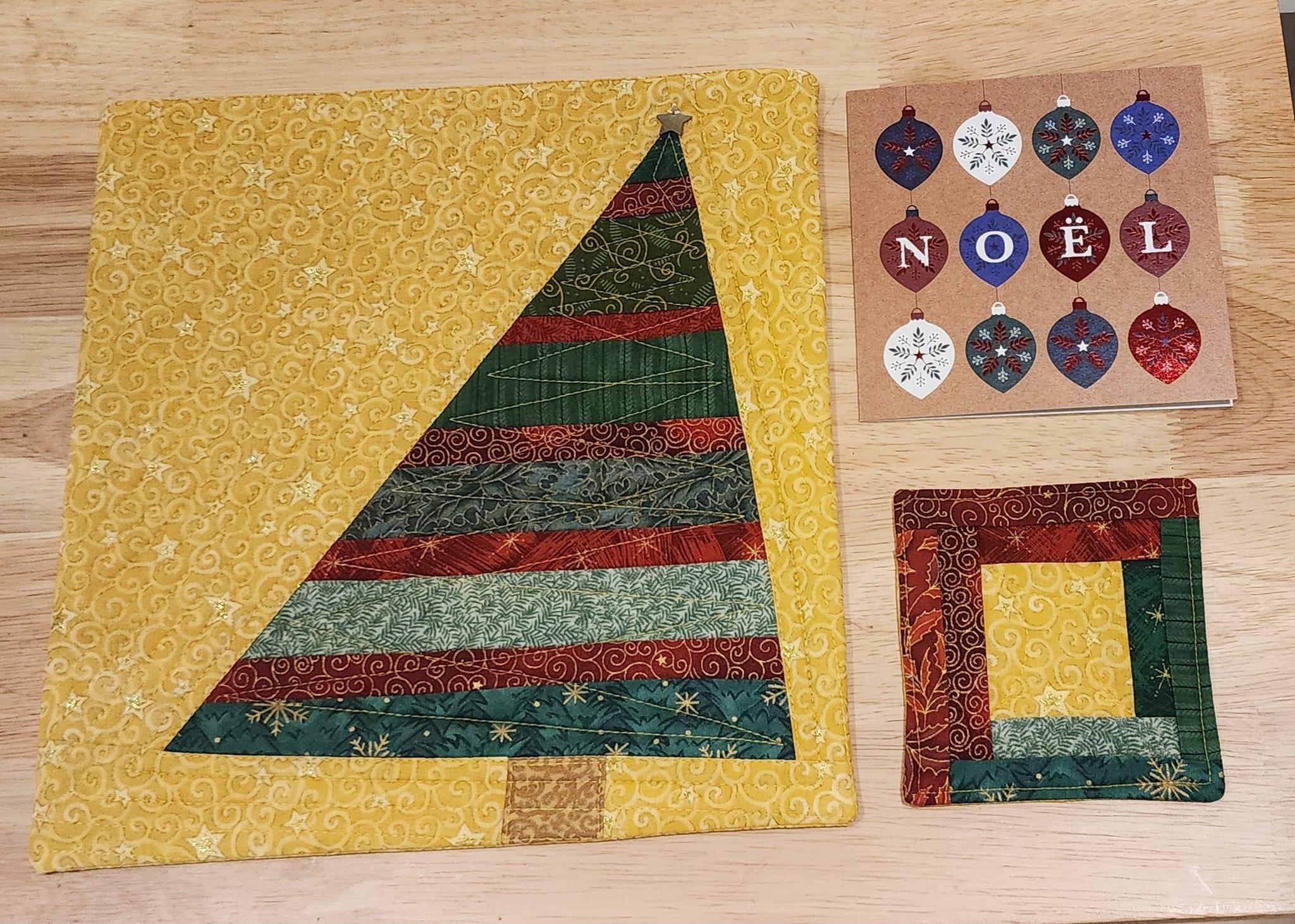 A Christmas themed placemat and coaster. The placemat features a strip pieced Christmas tree on a yellow swirl background. The coaster is made with a log cabin pattern with a yellow swirl square to match the background of the placemat and red and green fabric on the outer edges. A Christmas card with Noel and Christmas baubles is in the top right corner.