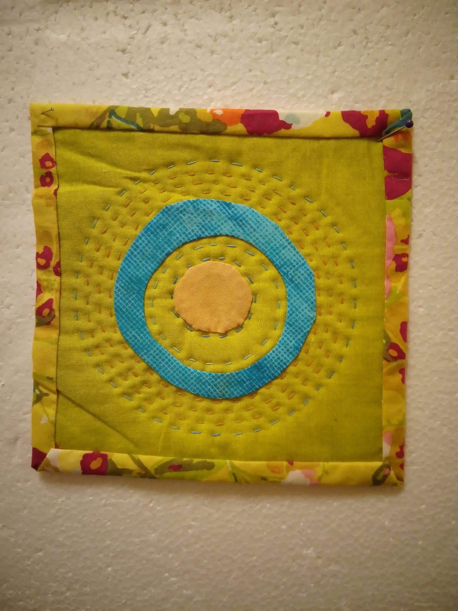 Lime green coaster with a central motif of a circle within a ring. Big hand stitching radiates out from the centre and the binding is a yellow floral fabric.
