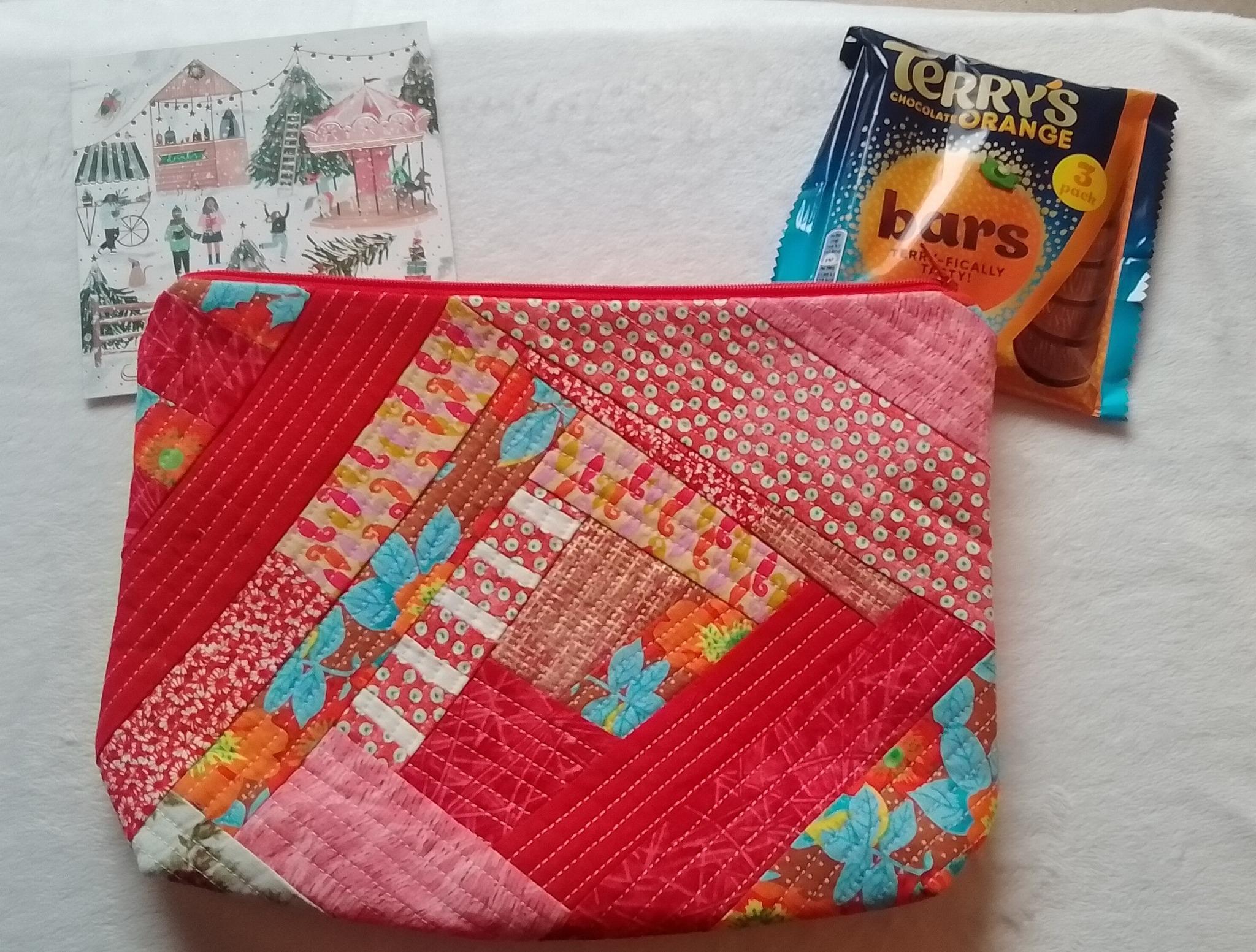 A fabric zippered pouch in red tones, with some accented blue florals. The pattern is log cabin inspired, radiating out from a central piece. In the background are a packet of Terry's chocolate orange bars and a Christmas Card featuring a market scene.