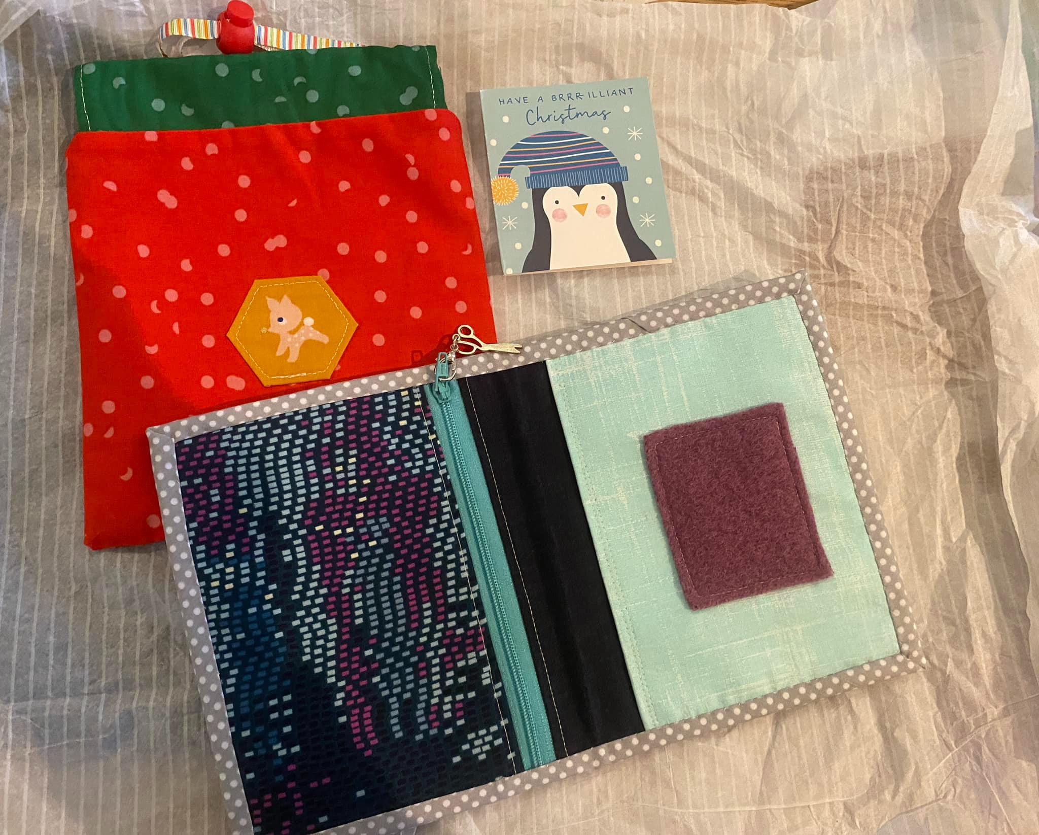 Zippered needlecase with a scissor zipper charm and a drawstring pouch made in Christmas colours with a reindeer hexagon feature in the centre. A Christmas card with a penguin is off to the right