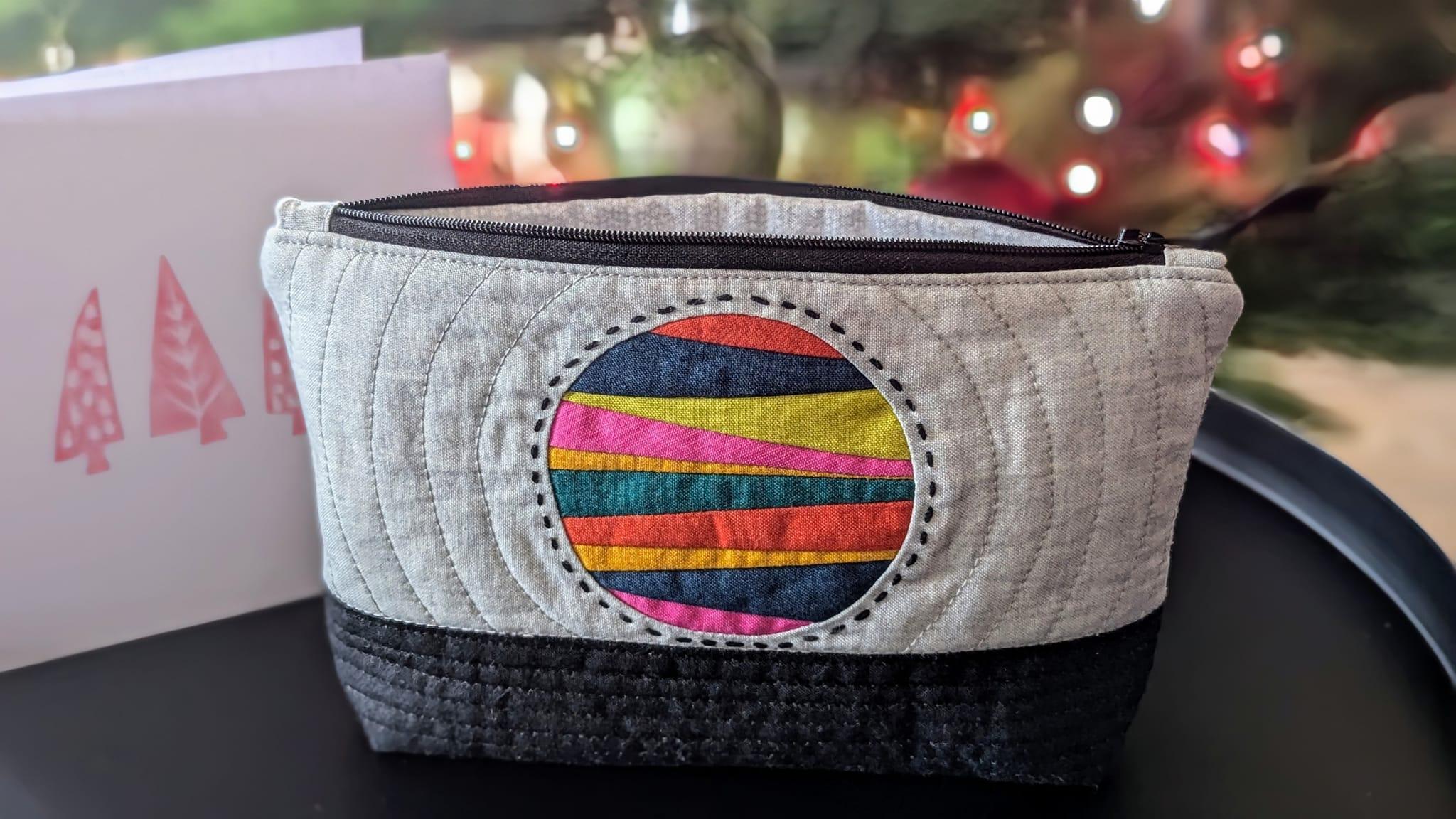 A white zippered pouch with a black bottom and a patchwork circle in the centre made of bright modern colours. The base of the bag is straight line stitched and the main section of the bag has circles radiating out from the circle. The circle has big hand stitching around it.