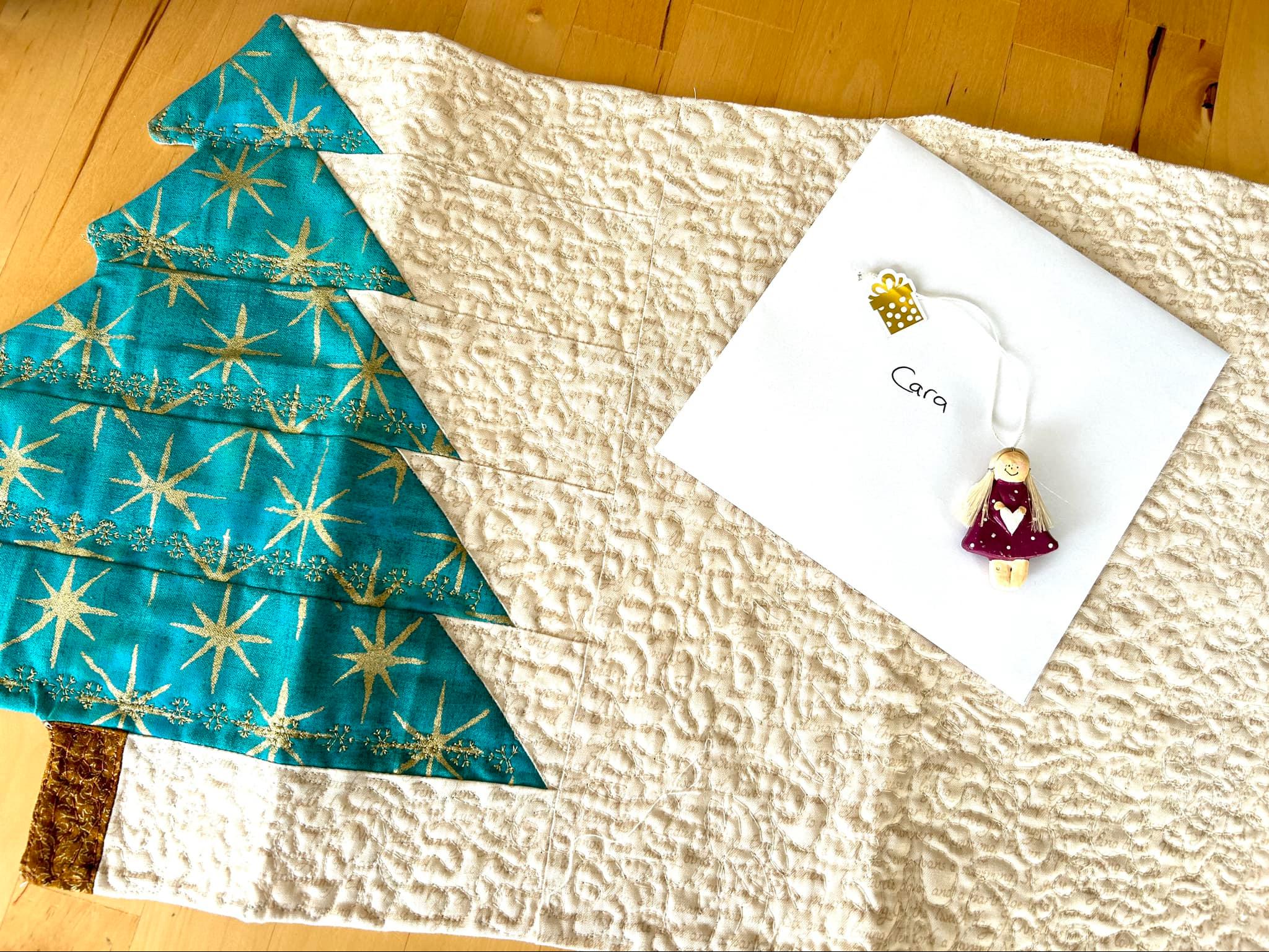 A cream placemat with a tree on one edge made of green and gold star fabric. A Card and ornament are placed on the placemat.