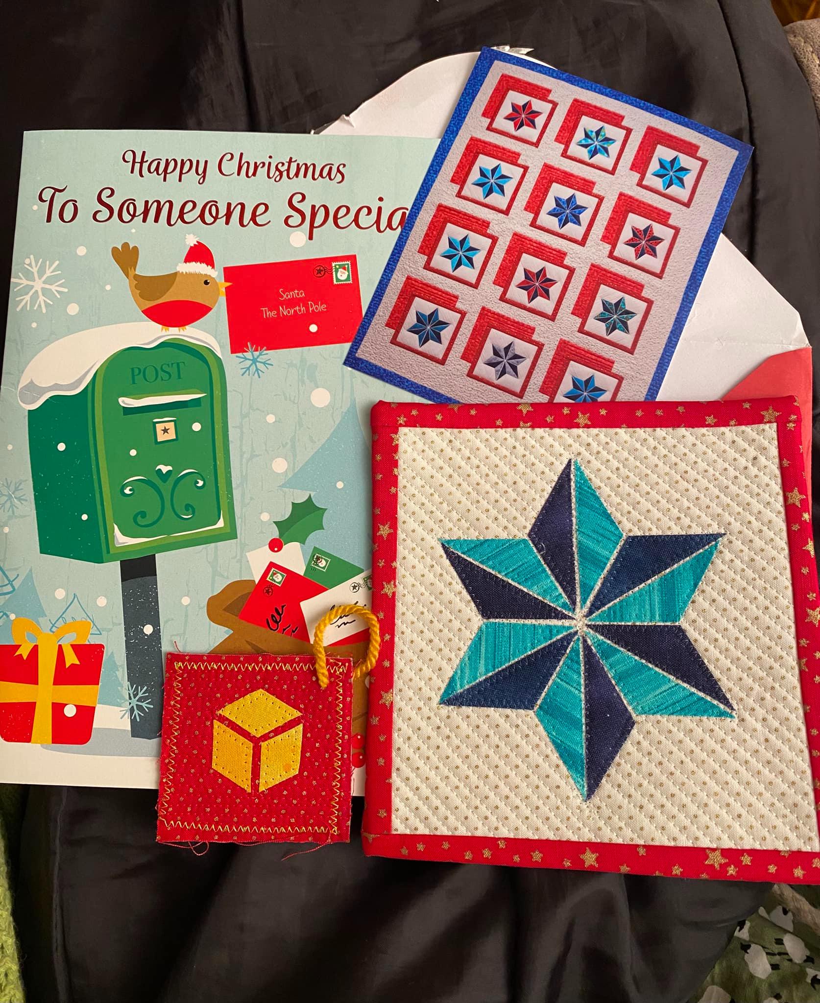 Mini Quilt of a blue start in light and dark blue. A white background is quilted in a radiating pattern out from the star and the mini quilt is bound with a red and gold star fabric. There is also a stitched gift tag.