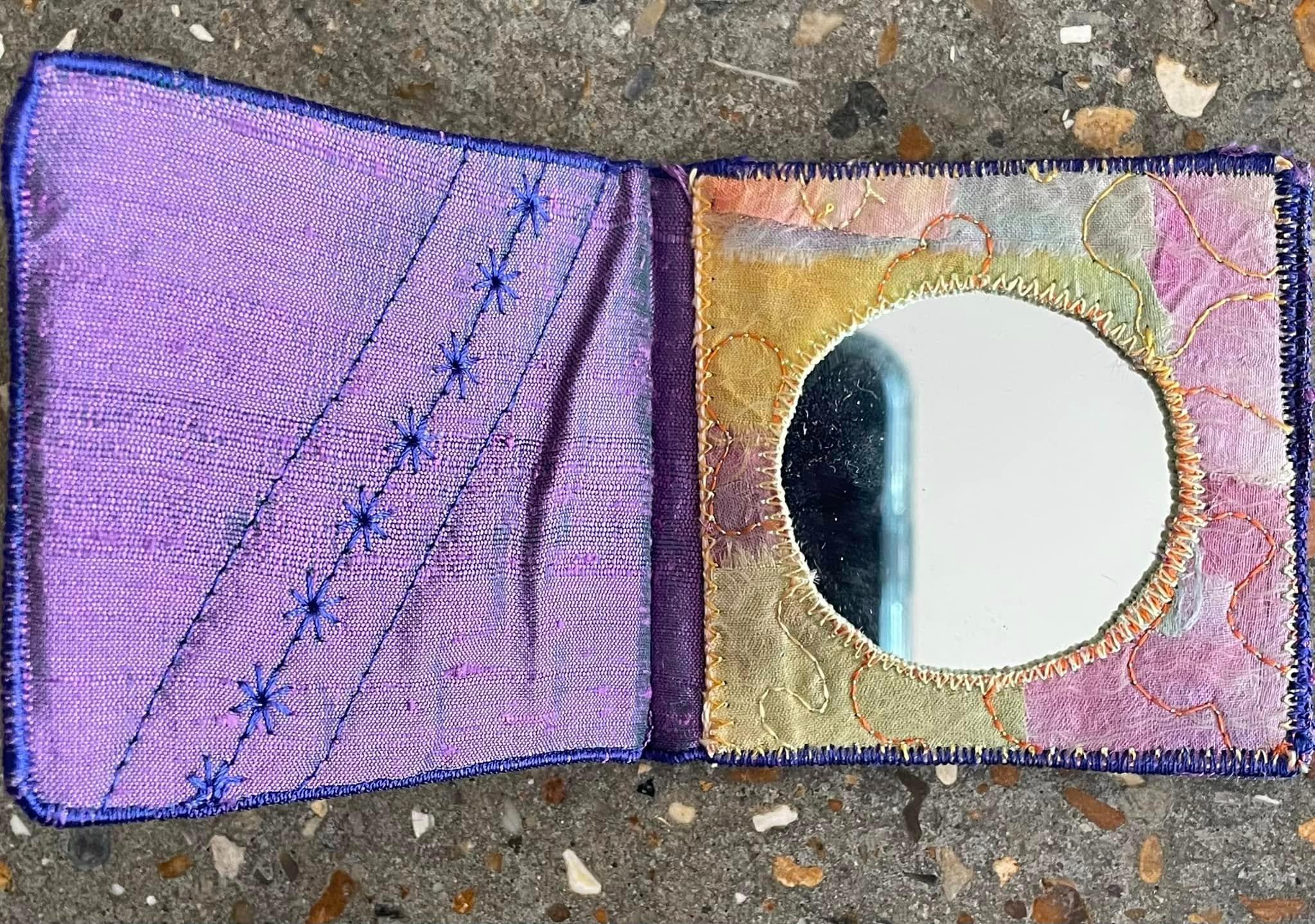 Compact Mirror case made from purple and some pink and yellow fabrics. The cover of the mirror has decorative star stitching running diagonally across it. A round mirror is encased in stitching on the inside of the cover.