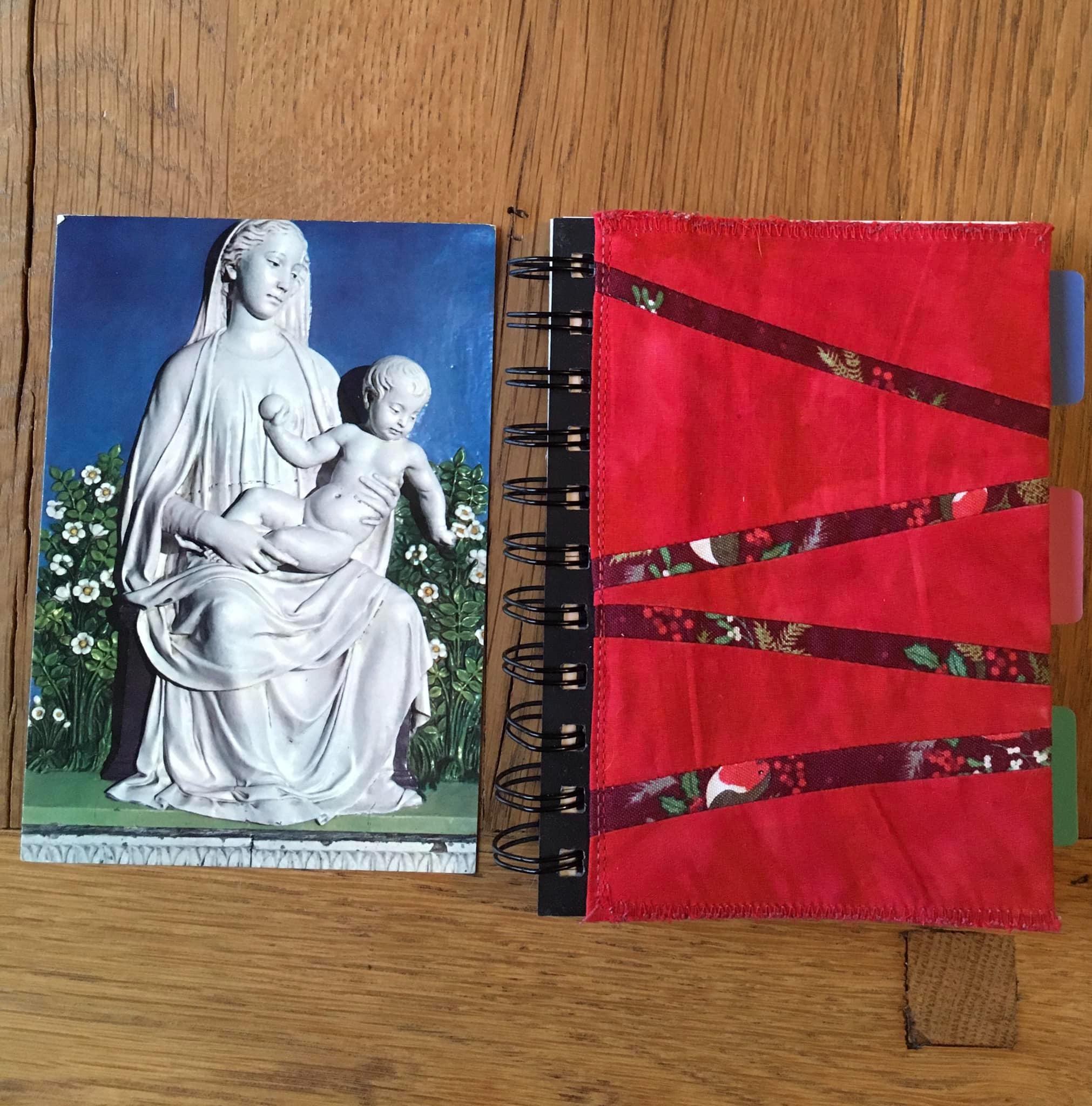 A notebook cover with a red background and skinny strips stretching across the cover horizontally in diagonal lines. The strips are made of Christmas fabric, with robins peeking through. To the left of the notebook cover is a postcard showing Mary and Jesus as a baby.