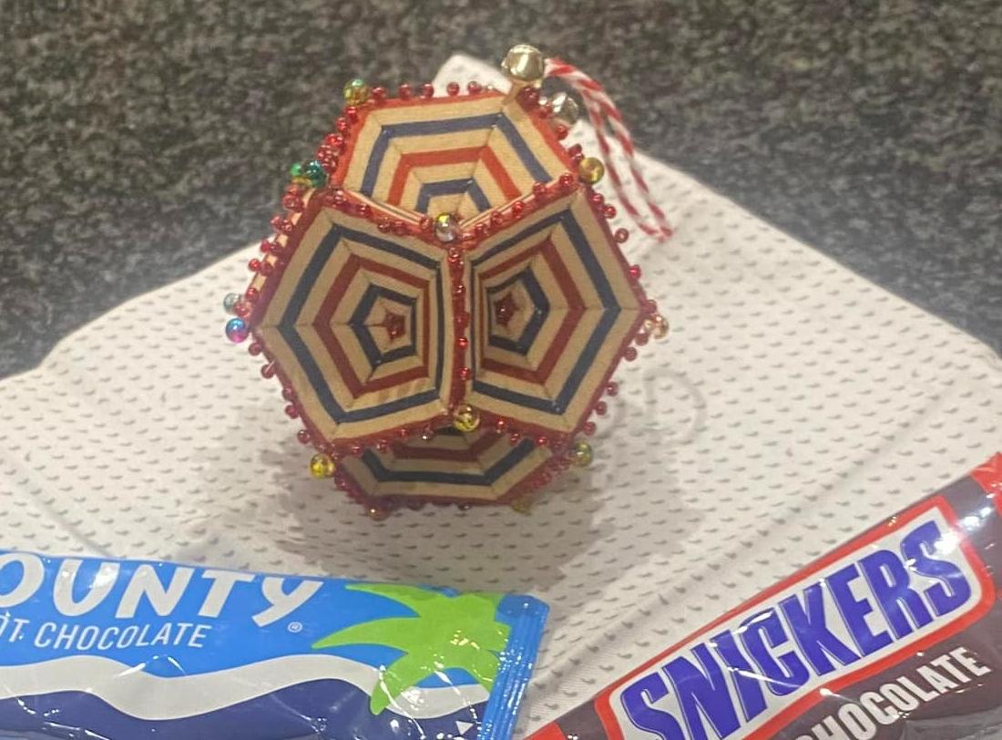 A beaded Christmas decoration. The decoration has concentric pentagons in red and blue radiating out of the centre of each side. Each side is pentagon shaped. In the foreground are 2 sachets of chocolate bar hot chocolates and the decoration sits on top of a fat quarter of fabric.