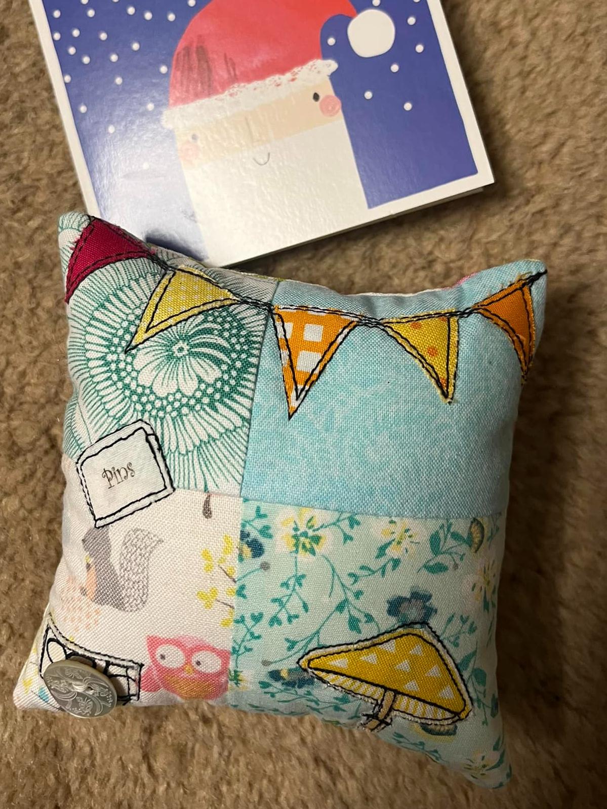 A patchwork pieced square pincushion with a line of bunting appliquéd and stitched at the top and an appliqué mushroom in the bottom right corner. In the bottom left corner is a silver button.
