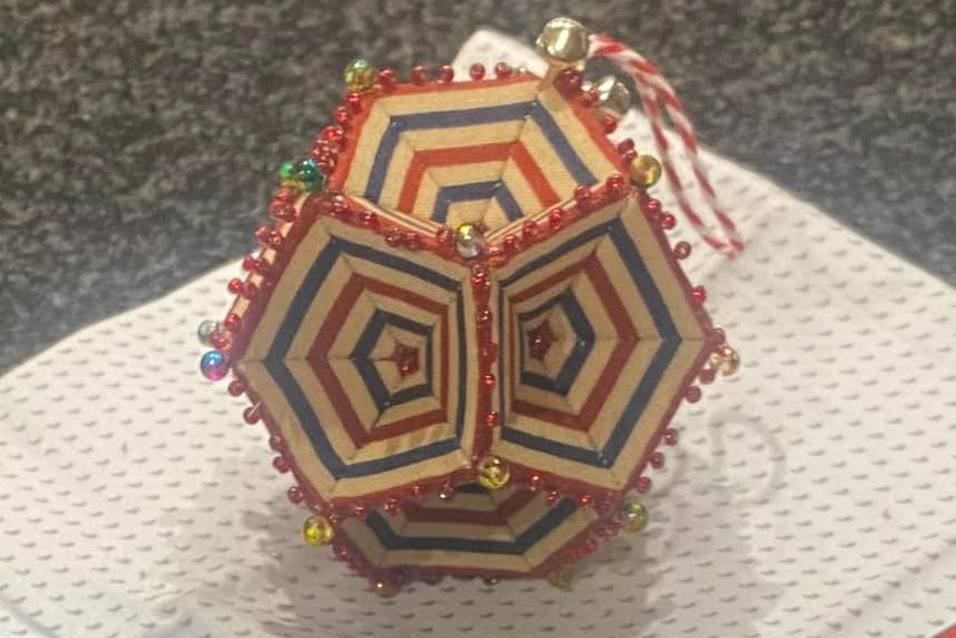 A beaded Christmas decoration. The decoration has concentric pentagons in red and blue radiating out of the centre of each side. Each side is pentagon shaped. In the foreground are 2 sachets of chocolate bar hot chocolates and the decoration sits on top o