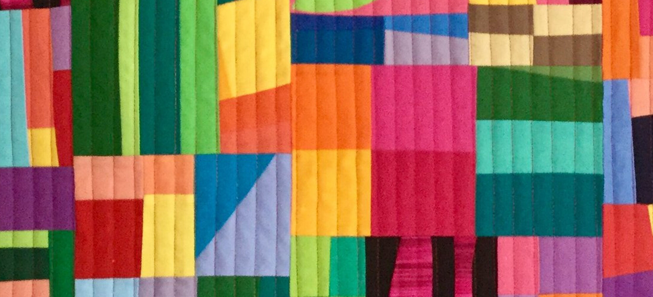 A colourful quilt with a patchwork design. The quilt is made up of various shapes, including rectangles, triangles, and squares, in vibrant colours like red, orange, yellow, green, blue, purple, and black. The shapes are arranged in a seemingly random pattern, creating a dynamic and abstract composition. The background is quilted with vertical lines.