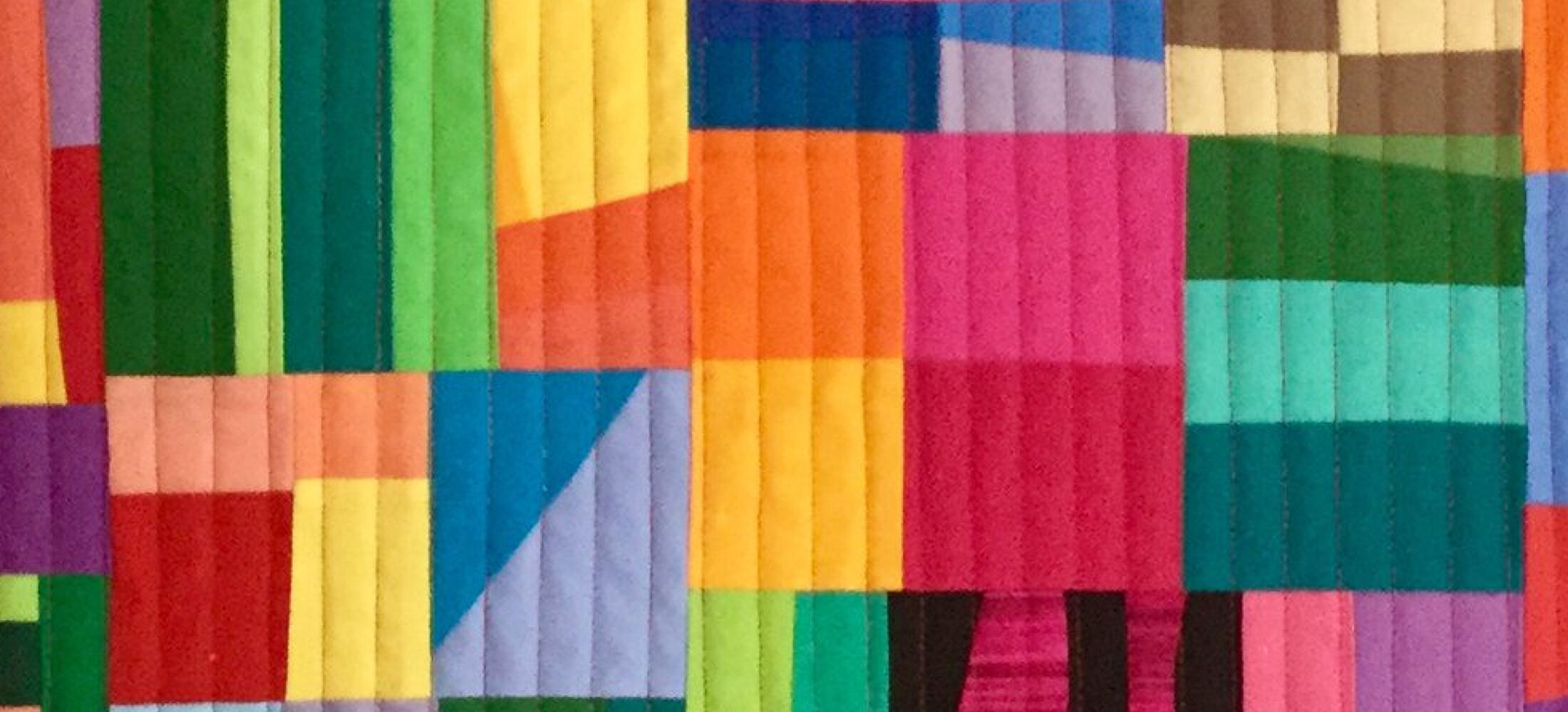 A colourful quilt with a patchwork design. The quilt is made up of various shapes, including rectangles, triangles, and squares, in vibrant colours like red, orange, yellow, green, blue, purple, and black. The shapes are arranged in a seemingly random pattern, creating a dynamic and abstract composition. The background is quilted with vertical lines.