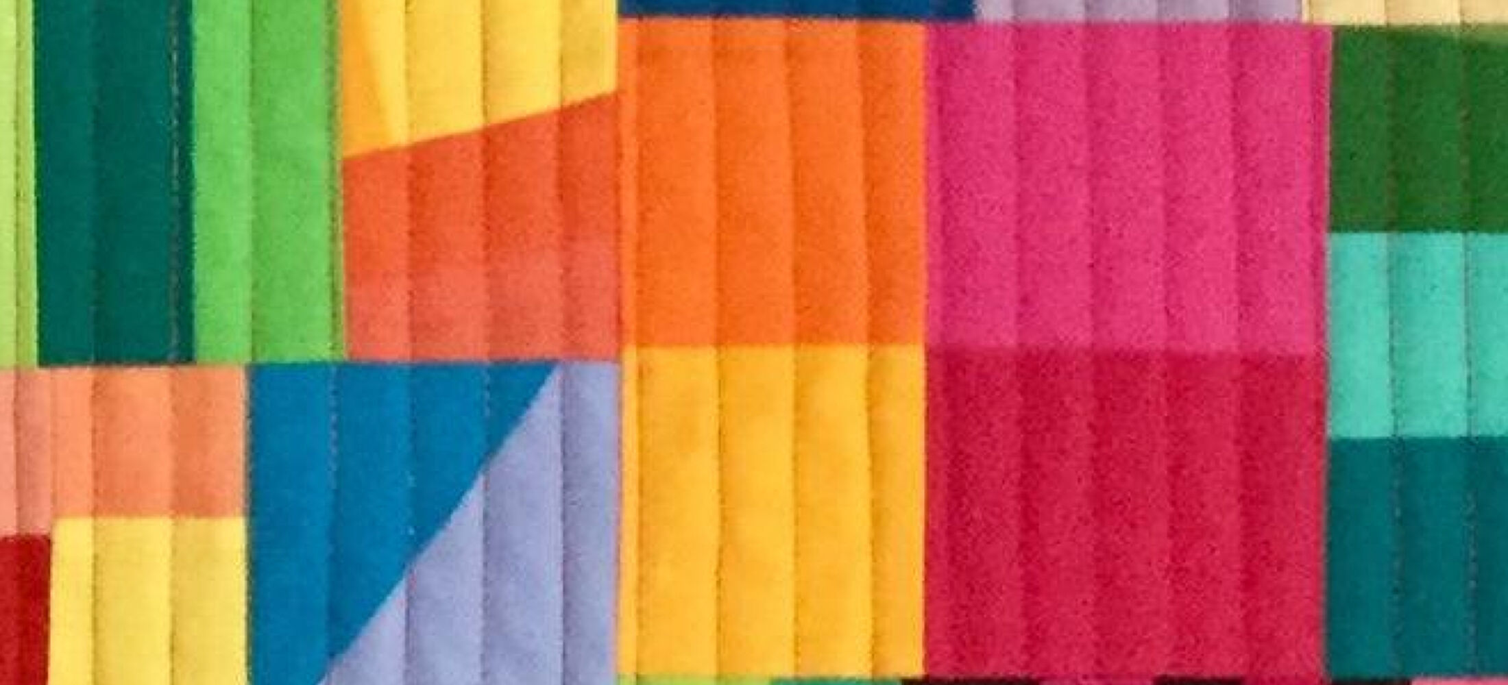 A colourful quilt with a patchwork design. The quilt is made up of various shapes, including rectangles, triangles, and squares, in vibrant colours like red, orange, yellow, green, blue, purple, and black. The shapes are arranged in a seemingly random pattern, creating a dynamic and abstract composition. The background is quilted with vertical lines.