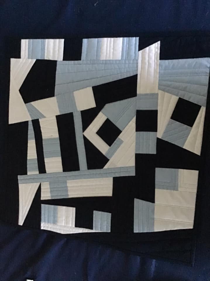A square quilt with a black background. The quilt features a geometric design of white and light blue shapes, including squares, triangles, and rectangles. The shapes are arranged in a seemingly random pattern, creating a dynamic and abstract composition. The background is quilted with a subtle, linear pattern.