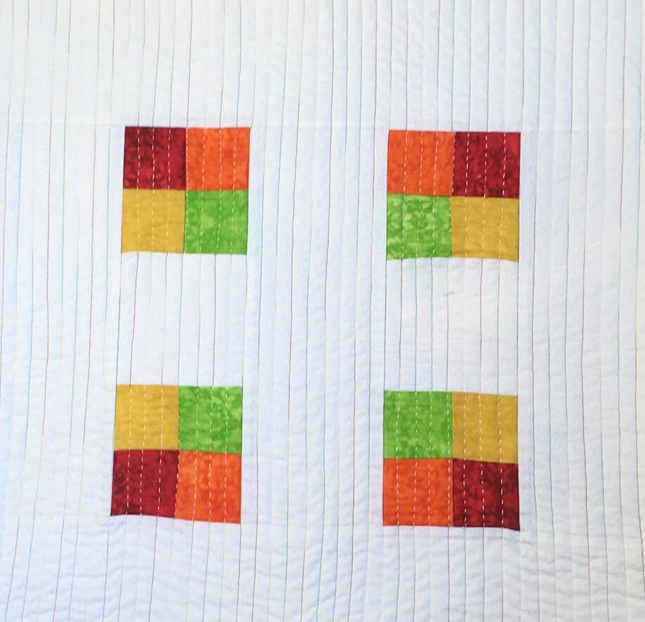 A small, rectangular quilt with a white background. The quilt features four squares arranged in two vertical pairs. Each square is made up of smaller squares in red, orange, yellow, and green. The background is quilted with vertical lines.