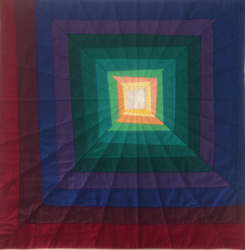 A square quilt with a colourful, geometric design. The quilt features a series of nested squares, each outlined in a different colour, creating a tunnel-like effect. The colours transition from dark red to purple, blue, green, yellow, and finally a light beige in the centre.