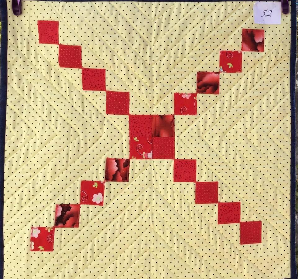 A small, square quilt with a yellow background. The quilt features a pattern of red squares arranged in a diagonal line, creating an "X" shape. The background fabric has a subtle, dotted pattern.