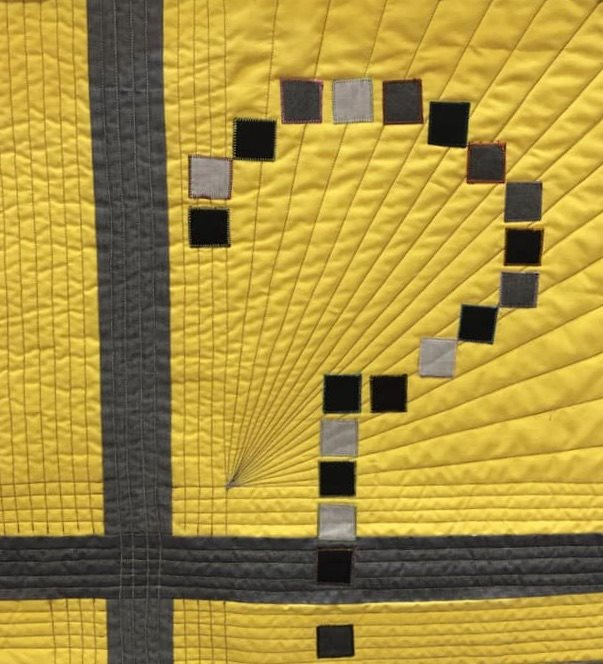 A close-up of a quilt with a bright yellow background. The quilt features a large, stylized question mark made up of black and grey squares. The question mark is surrounded by radiating lines of stitching.