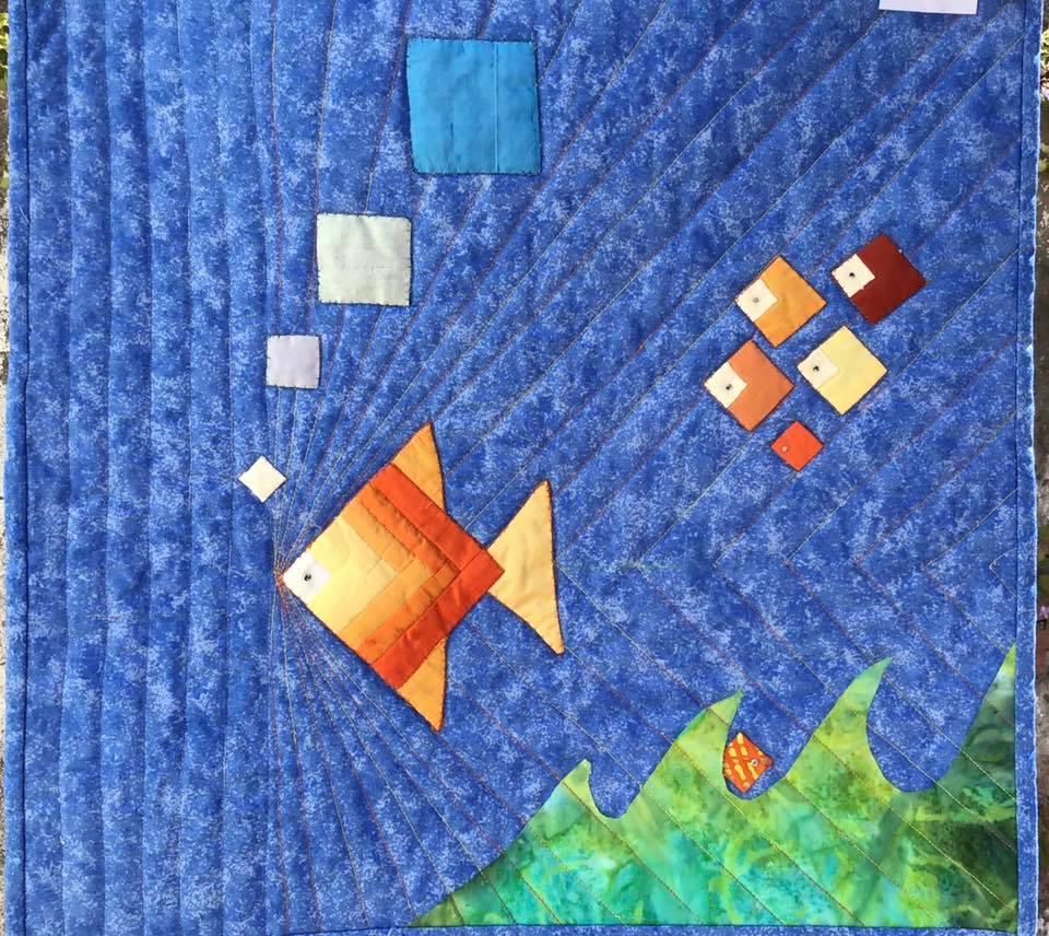 A small, square quilt with a blue background depicting an underwater scene. A large orange fish swims towards the top left corner, surrounded by smaller orange shapes representing bubbles. The bottom of the quilt shows a green area with waves, suggesting the ocean floor.