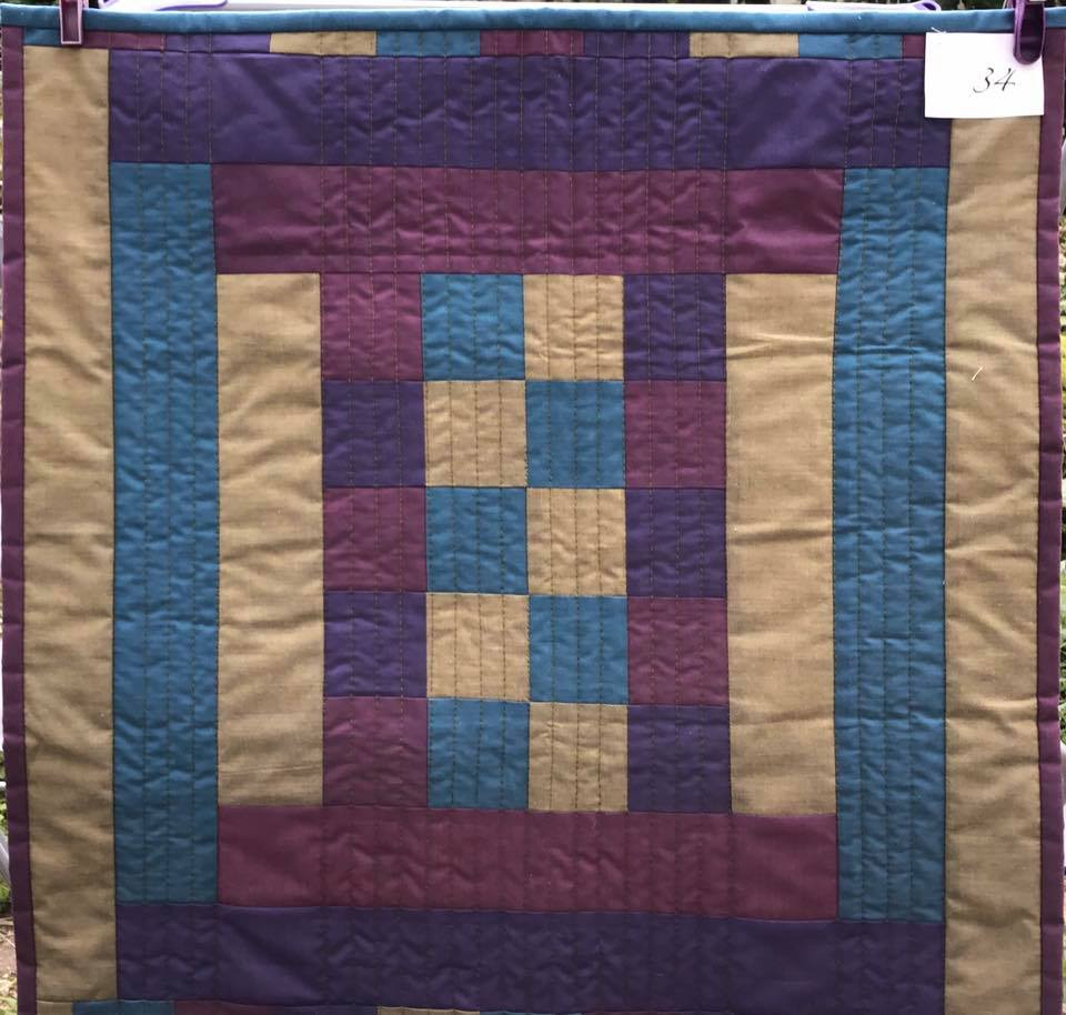 A small, square quilt with a patchwork design. It features a combination of purple, blue, and beige fabrics arranged in a grid-like pattern. The quilt has a textured, quilted pattern.