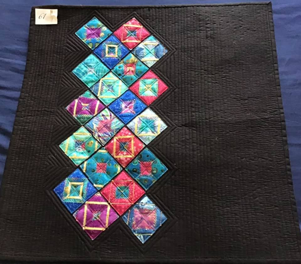 A small, square quilt with a black background. The quilt features a vertical line of colourful squares arranged in a diamond pattern. The squares are made of various fabrics with different patterns and textures, including some with metallic accents. The background is quilted with a subtle diamond pattern.