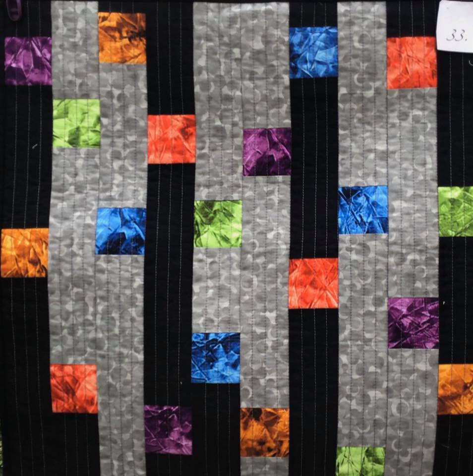 A small, rectangular quilt with a black background. The quilt features a pattern of colourful squares in various shades of orange, blue, purple, and green, arranged in a staggered pattern on a grey background.