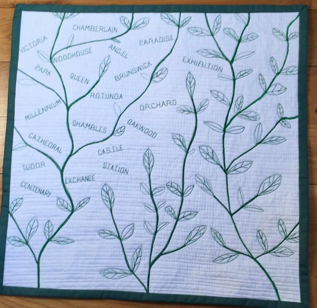 A quilt with a white background and a green border. The quilt features a tree-like design with branches and leaves. On each branch, there are embroidered names of streets, such as Cathedral, Tudor, Queen, and others.