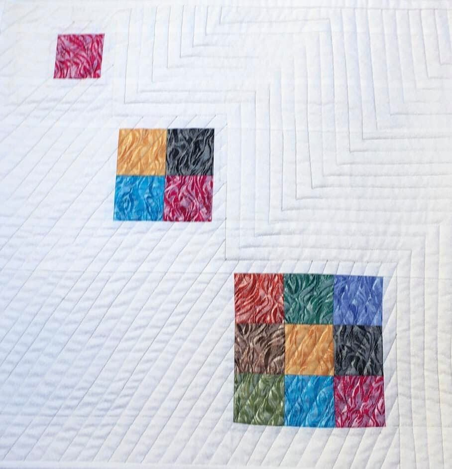 A quilt with a white background. The quilt features three clusters of colourful squares arranged in a diagonal line. Each cluster consists of smaller squares in various colours, including red, orange, yellow, blue, and green. The background is quilted with a pattern of diagonal lines.
