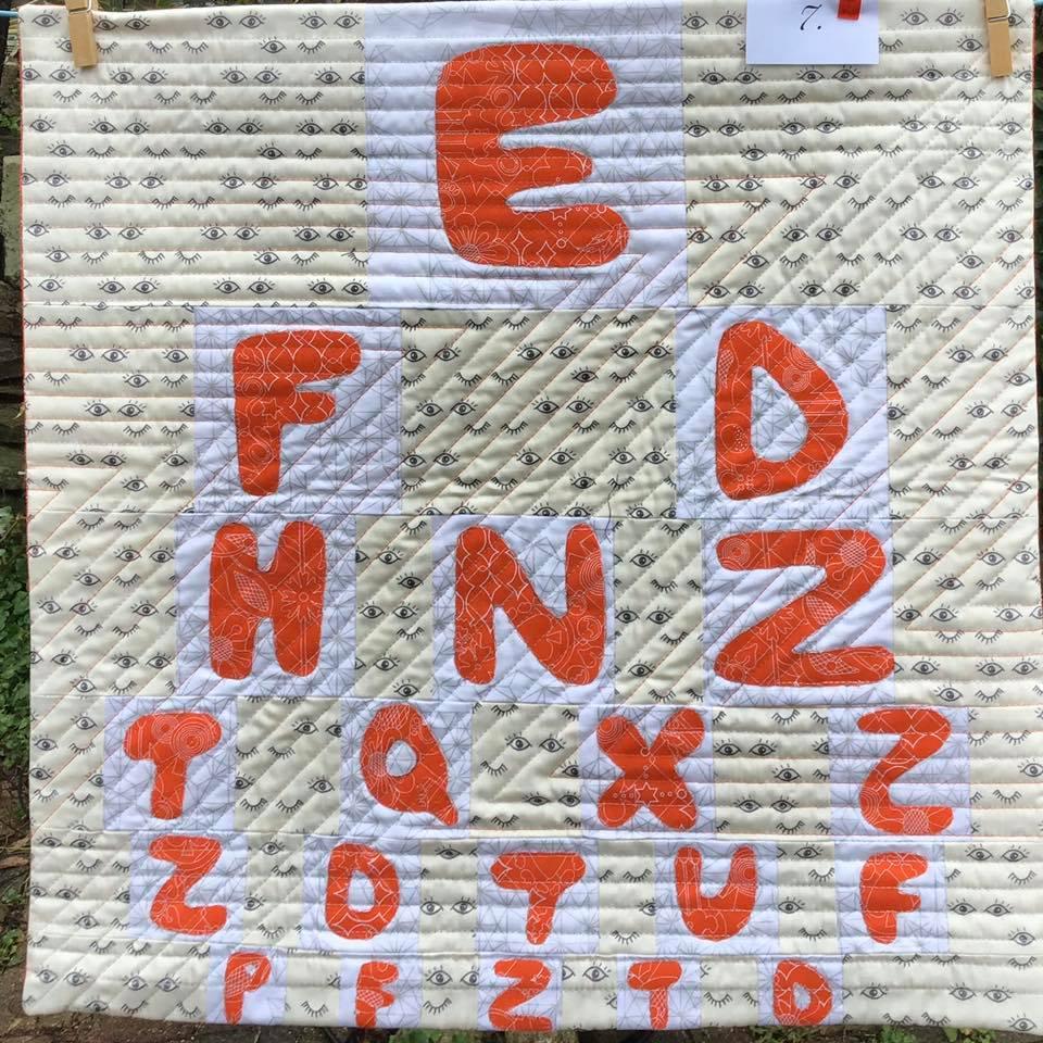 A small, square quilt with a beige background. The quilt features an eye chart pattern with the letters of the alphabet in orange, arranged in rows. The background fabric has a pattern of small, black eyes.