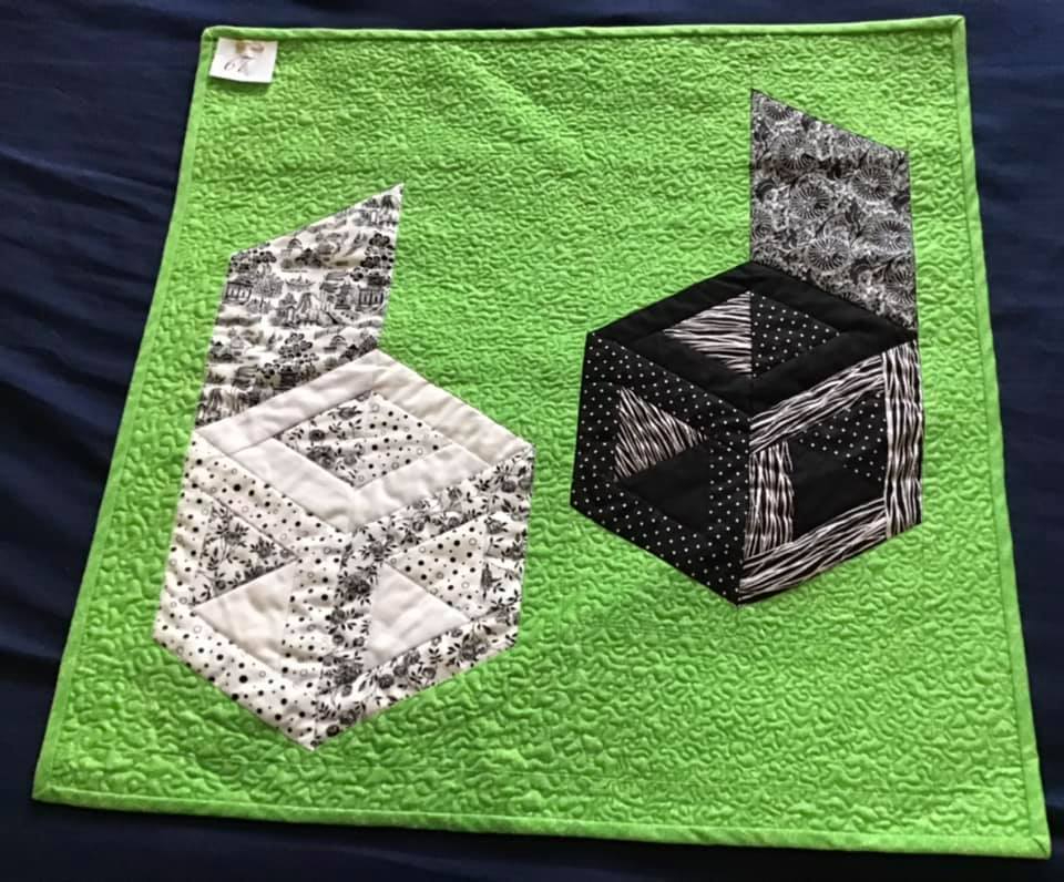 A square quilt with a green background. The quilt features two 3D-looking boxes made from black and white patterned fabric. One box is open, revealing a smaller box inside. The background is quilted with a textured pattern.
