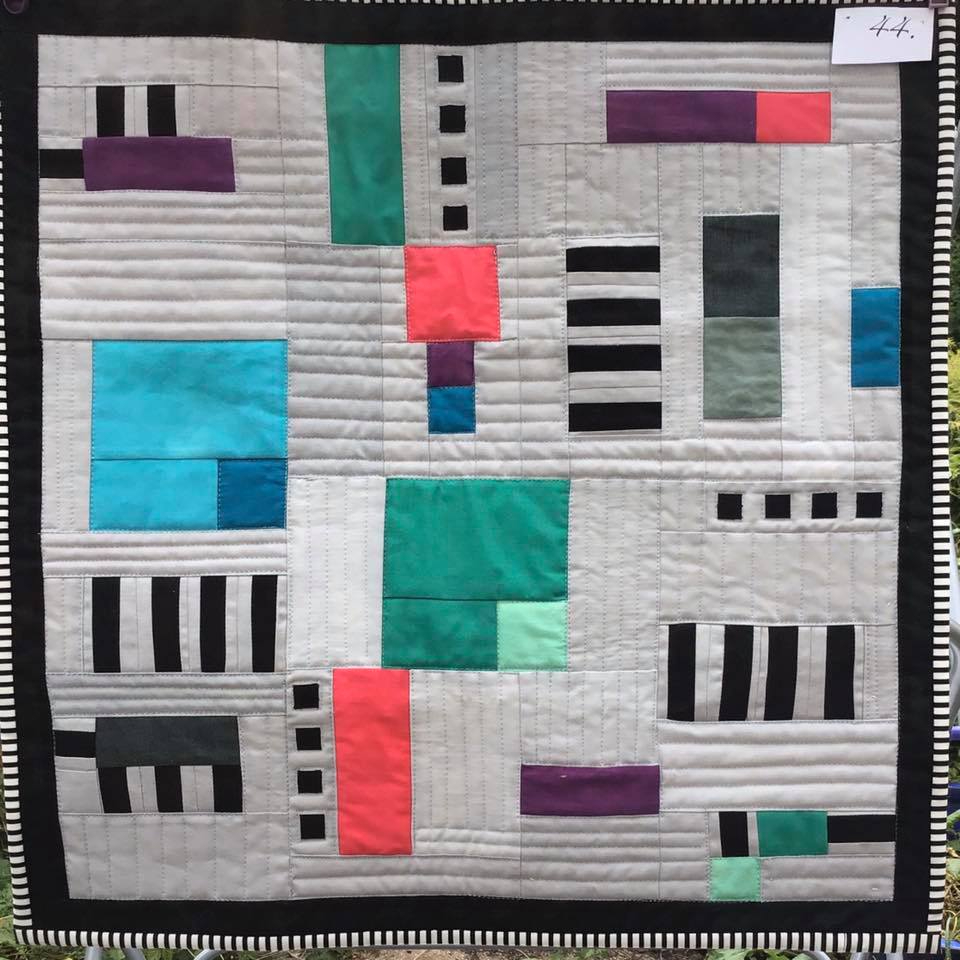 A small, square quilt with a patchwork design. It features a variety of colours and patterns, including black and white stripes, solid colours, and geometric shapes. The overall design is abstract and modern.
