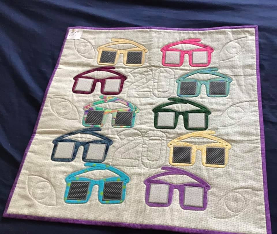 A colourful quilt with a playful design of glasses. Each pair of glasses has a different coloured frame and lenses, some with patterns or textures. The background is a light beige with a subtle pattern.