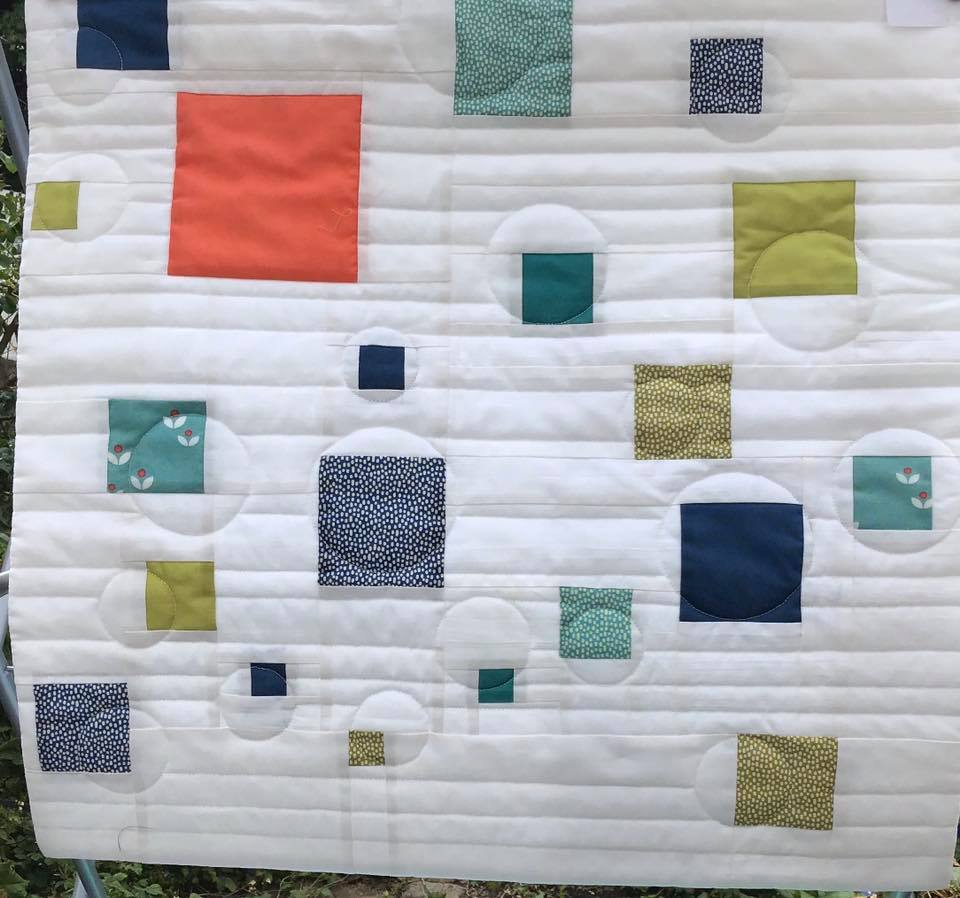 A small, square quilt with a white background. The quilt features a pattern of colourful squares and circles of various sizes, arranged in a seemingly random fashion. The background is quilted with vertical lines.