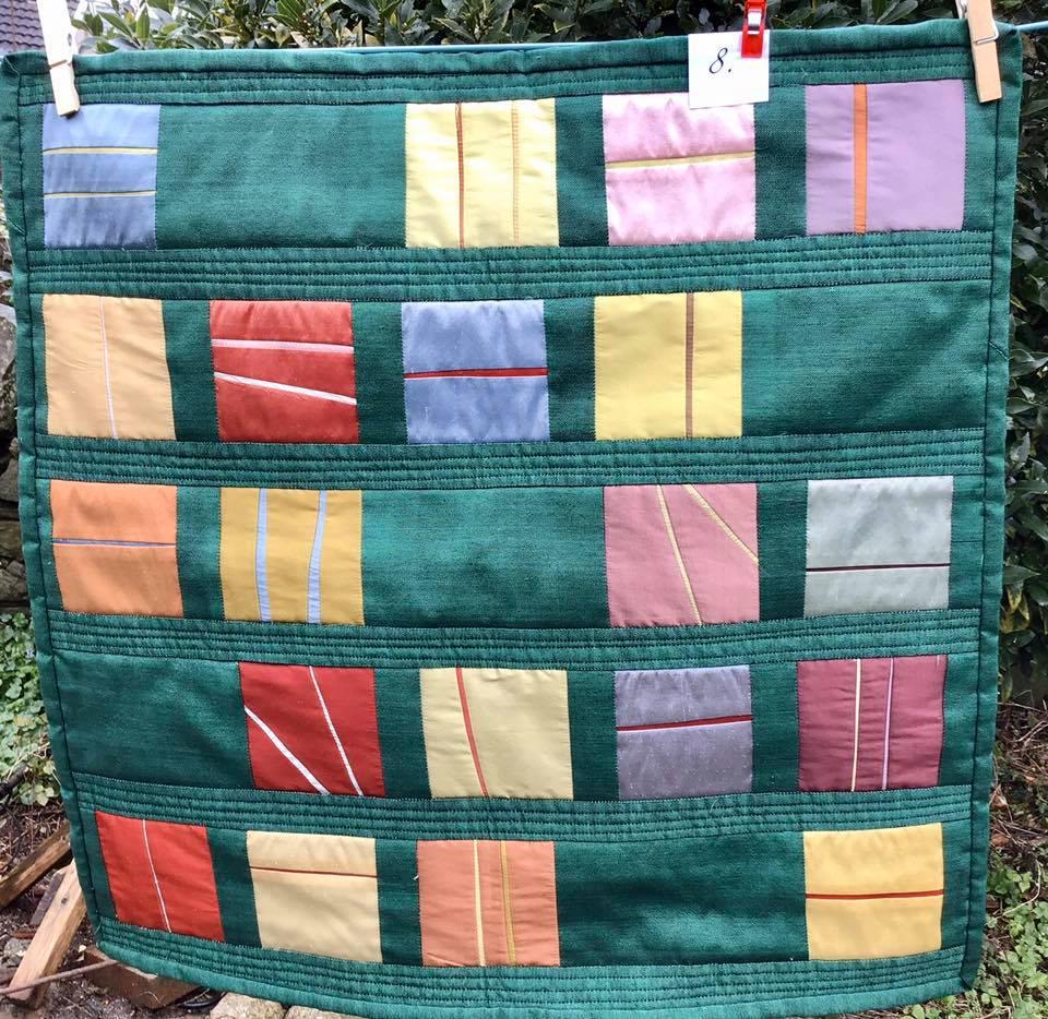 A small, square quilt with a green background. The quilt features a grid of rectangles in various colours, including pink, yellow, orange, and blue. Each rectangle has a thin, red line across it.