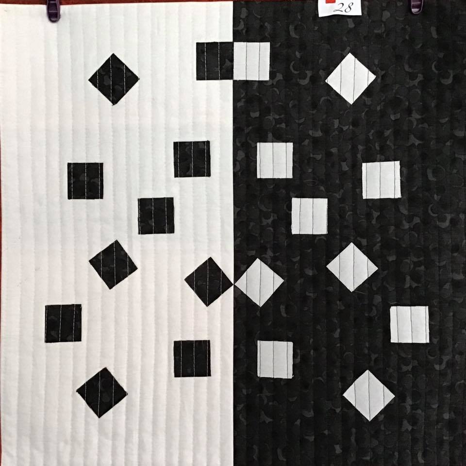 A small, square quilt with a black and white design. The quilt is divided into two halves, one black and one white. Each half features a pattern of squares and diamonds of the opposite colour.