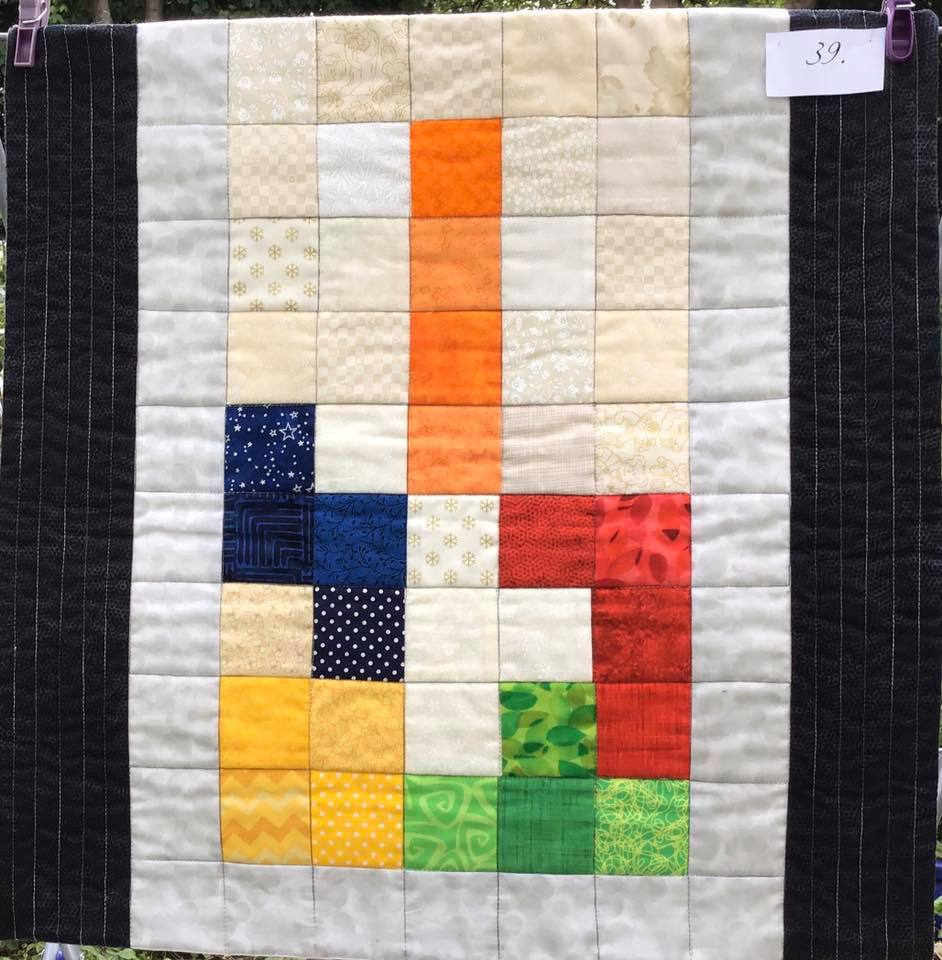A small, square quilt with a white background and black borders. The quilt features a colourful "L" shape made up of small squares in various colours, including orange, red, green, blue, and yellow.