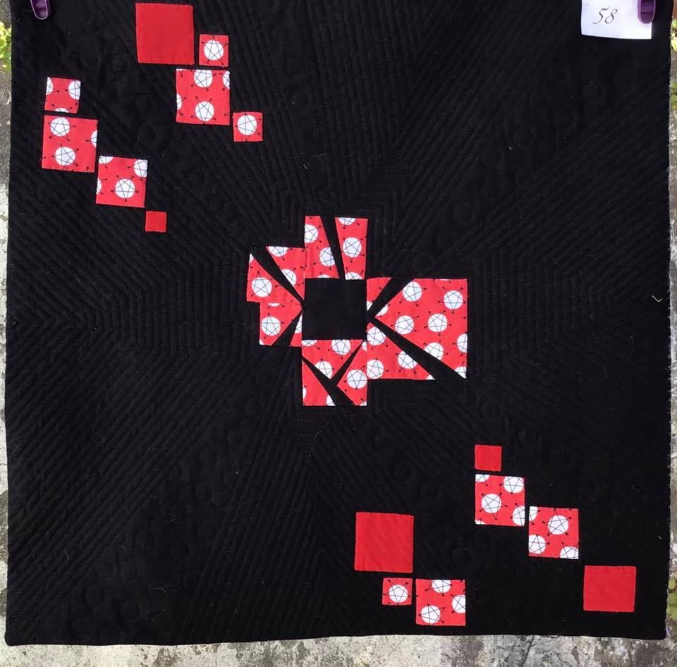 A small, square quilt with a black background. The quilt features a central design of red squares with white polka dots, arranged in a radial pattern. There are also smaller clusters of these squares in the corners of the quilt.