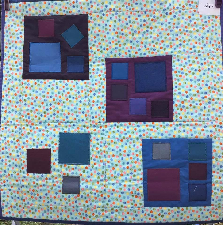 A small, square quilt with a blue background covered in colourful dots. The quilt features multiple squares of varying sizes and shades of blue and purple, arranged in groups.