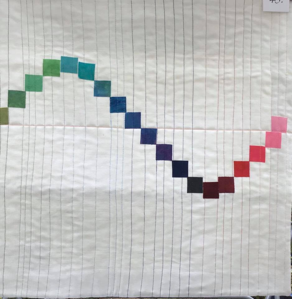 A small, square quilt with a white background. The quilt features a colourful, wavy line made up of small squares in various shades of green, blue, purple, and red. The background is quilted with vertical lines.