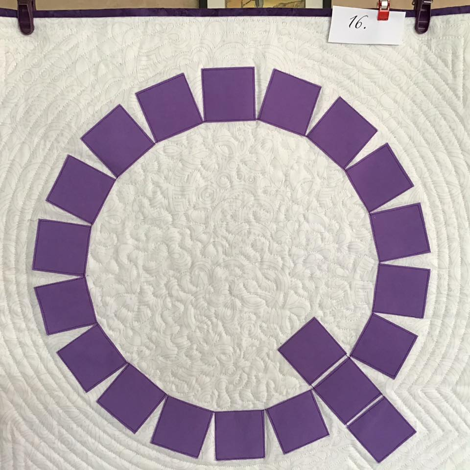 A small, square quilt with a white background. The quilt features a large, purple letter "Q" made up of individual squares. The background is quilted with a circular pattern.