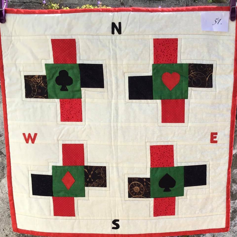A small, square quilt with a white background. The quilt features a compass design with the four cardinal directions (North, South, East, West) labeled. Each quadrant has a playing card suit symbol (spades, hearts, diamonds, clubs) in the centre. The background fabric has a subtle pattern of gears.