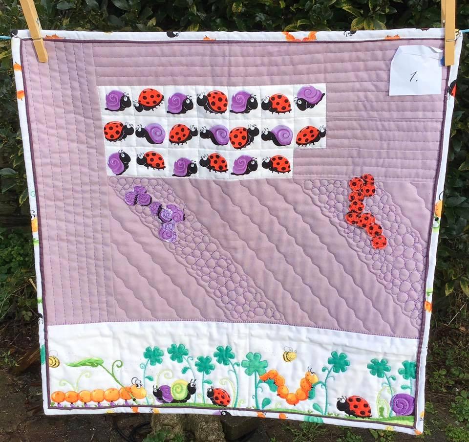 A square quilt with a lavender background. The quilt features a row of colourful ladybirds and snails at the top, and a row of caterpillars, ladybirds, and snails at the bottom. The background is quilted with a wavy pattern.
