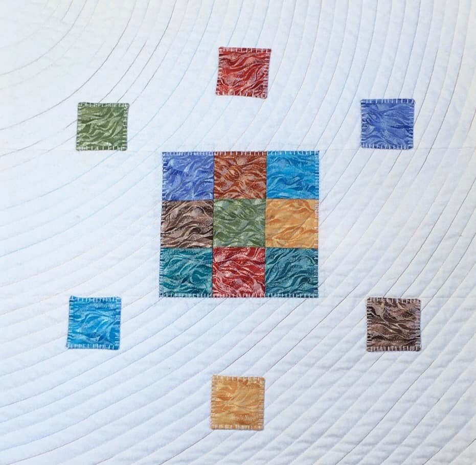 A small, square quilt with a white background. The quilt features a central square made up of smaller squares in various colours, including blue, green, brown, and orange. Surrounding the central square are additional squares of different colours, arranged in a circular pattern. The background is quilted with curved lines.