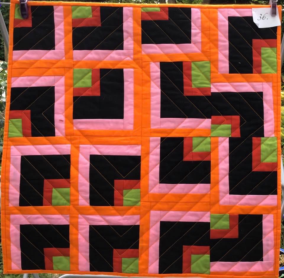 A small, square quilt with a patchwork design. It features a repeating pattern of squares within squares, using black, pink, green, and orange fabric. The background is quilted with a diamond pattern.